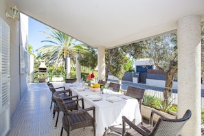 Een overdekt terras met eethoek voor vier personen, omringd door rieten stoelen, met zicht op palmbomen en een gebouw op de achtergrond.