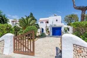 A white building with a blue door, surrounded by greenery and a stone pathway leading to the entrance.