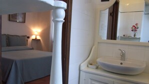 Una camera da letto con un letto, una lampada da comodino, un pilastro bianco, e un bagno con lavandino e specchio.