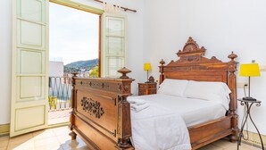 A wooden four-poster bed with white linens, a nightstand with a lamp, and a balcony with a view of the city.