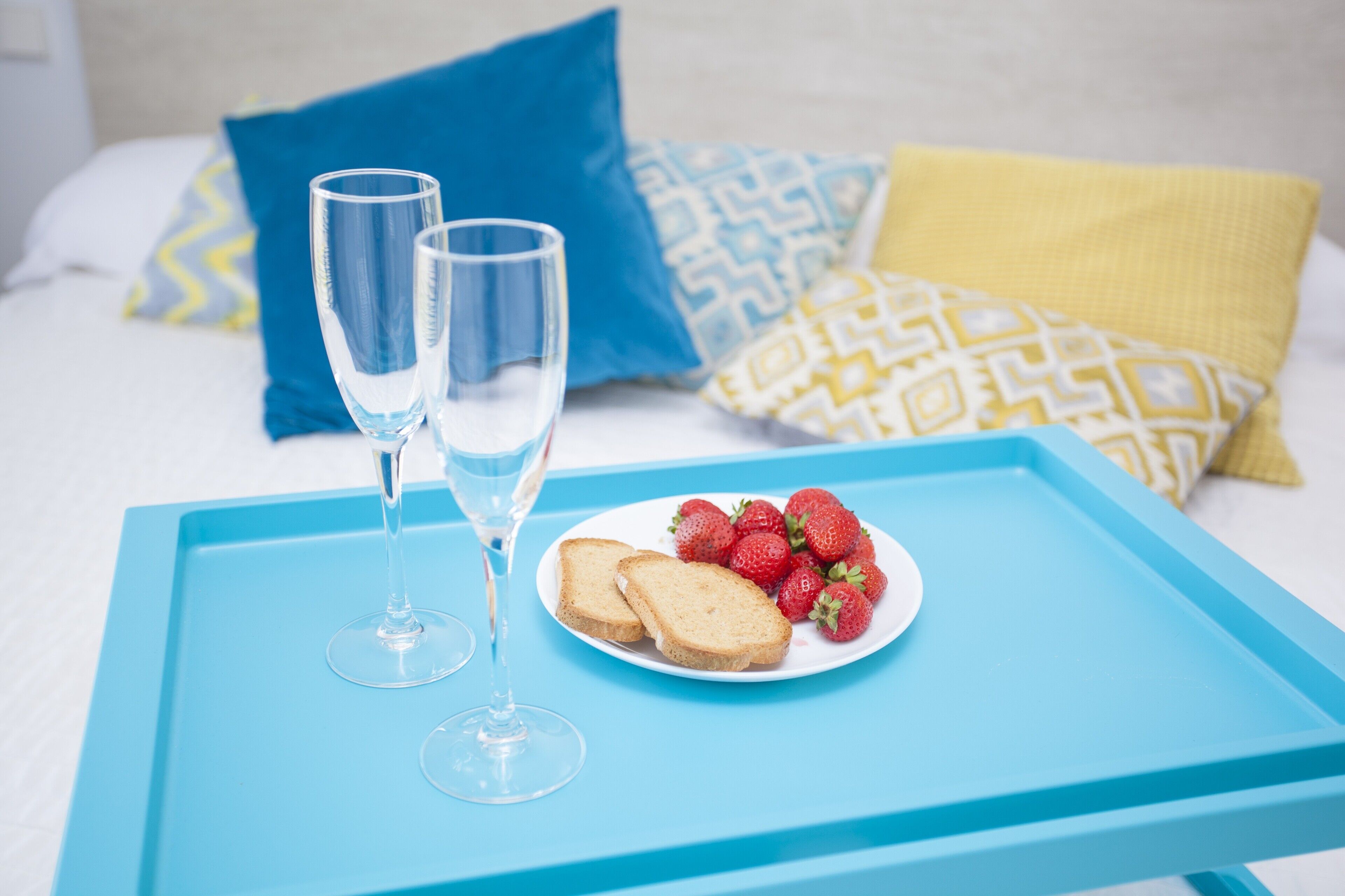 A bed with blue and yellow pillows, a tray with two glasses and a plate of strawberries and toast.