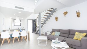 Ein moderner Wohnraum mit Essbereich, einem grauen Sofa mit bunten Kissen, einem weißen Couchtisch und einer Treppe mit individuellem Design.