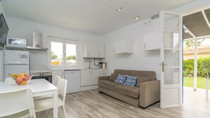 Una cocina compacta con electrodomésticos blancos, un comedor con mesa y sillas, un sofá color beige con un cojín estampado y un puerta corrediza de vidrio que da a un espacio exterior.
