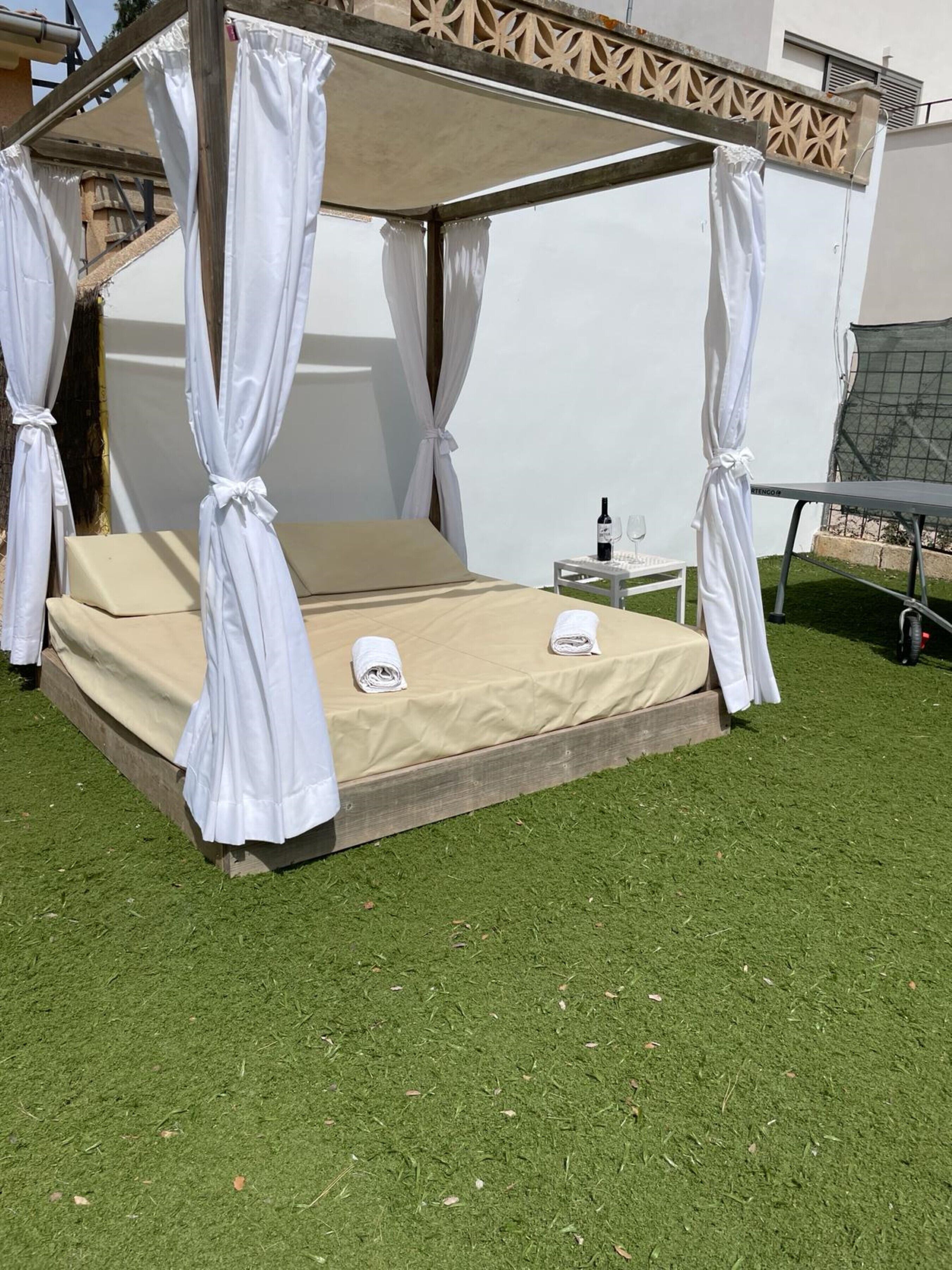 Un lit à baldaquin avec des rideaux blancs, un matelas beige et une petite table sur laquelle se trouvent une bouteille et des verres.