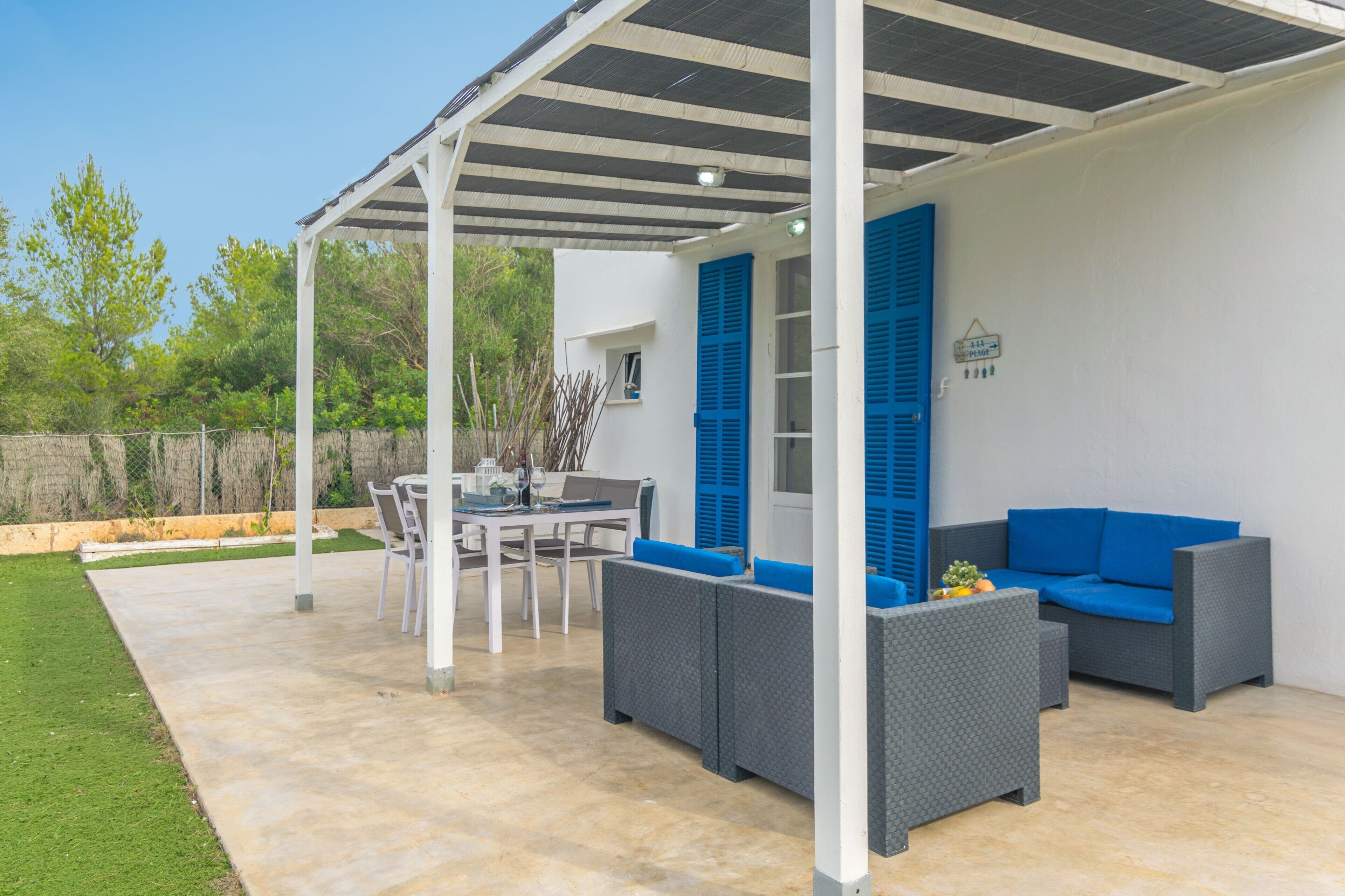 Un patio al aire libre con una zona de estar cubierta, una mesa de comedor y un edificio blanco con contraventanas azules.