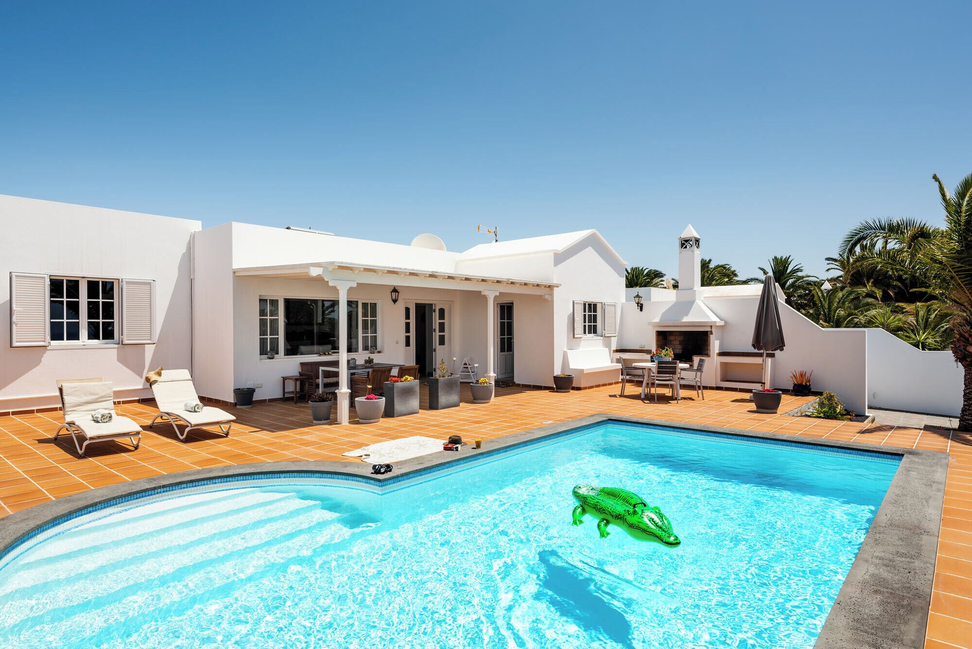 Una piscina con un juguete inflable verde, un porche cubierto con mobiliario exterior y un edificio blanco con persianas.