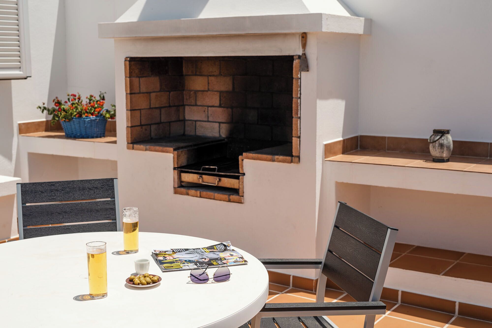 Un patio al aire libre con una parrilla de ladrillo, una mesa redonda con una bebida, una revista y un cuenco de aceitunas.