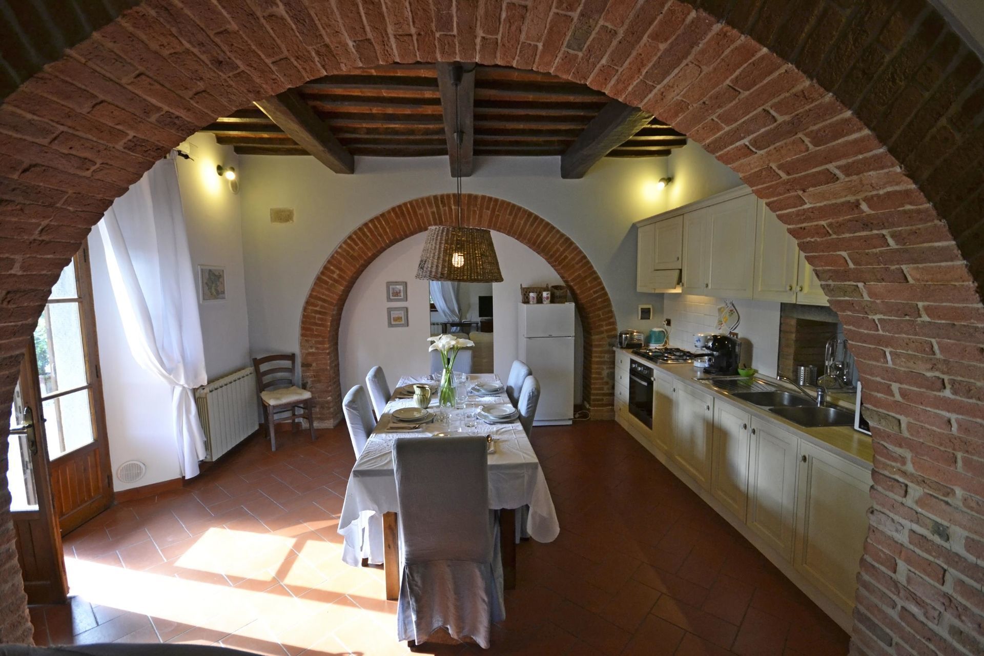 Un coin repas agrémenté d’une arche en briques, une table dressée pour quatre personnes, et une cuisine avec des meubles blancs et un fourneau.