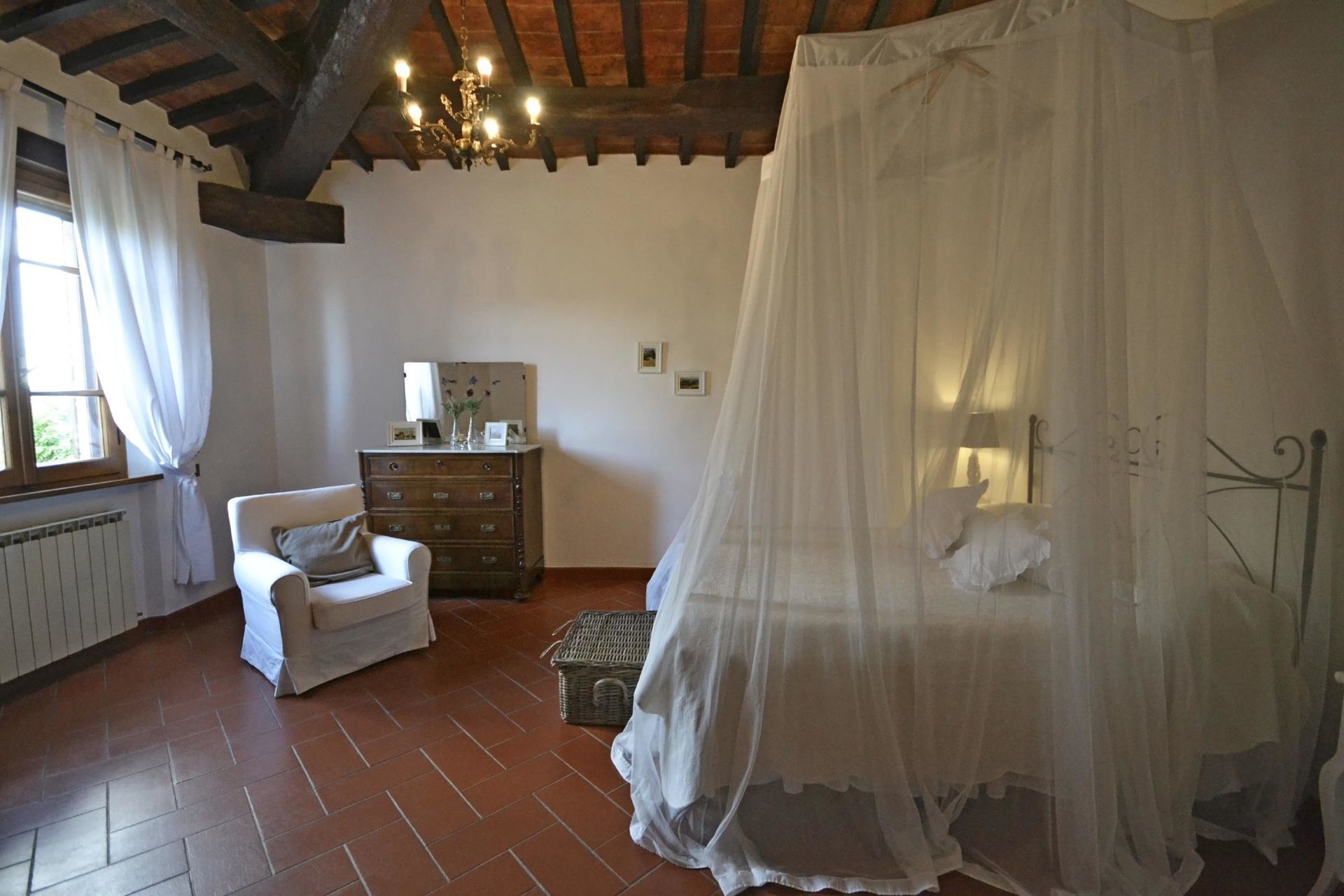 A bedroom with a bed covered by a white canopy, a wooden dresser, a white chair, and a window with curtains.