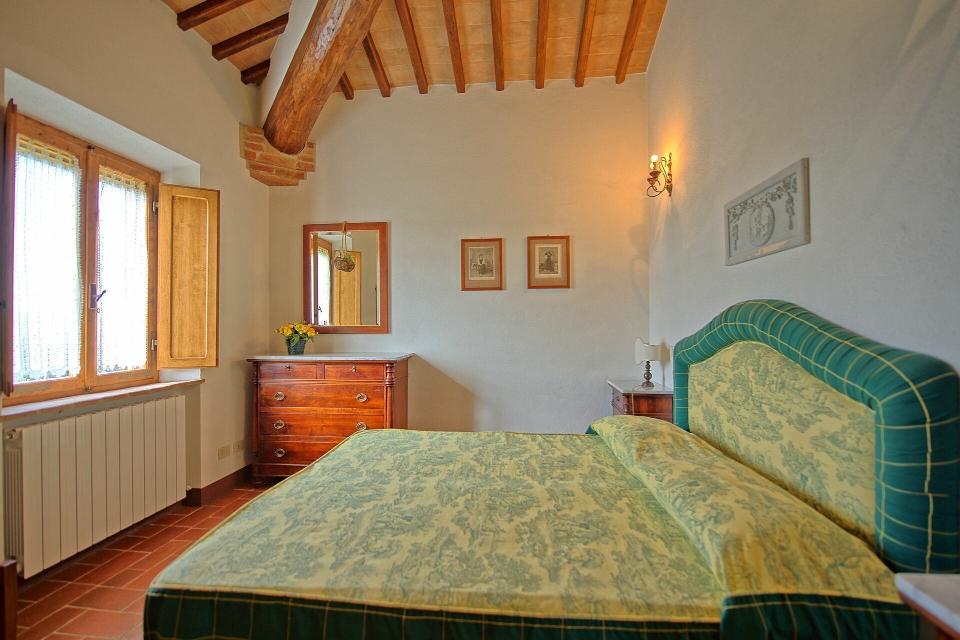 Une chambre avec un plafond en bois, un lit recouvert d’une couvre-lit à motifs, une commode, un miroir et deux tableaux encadrés accrochés au mur.