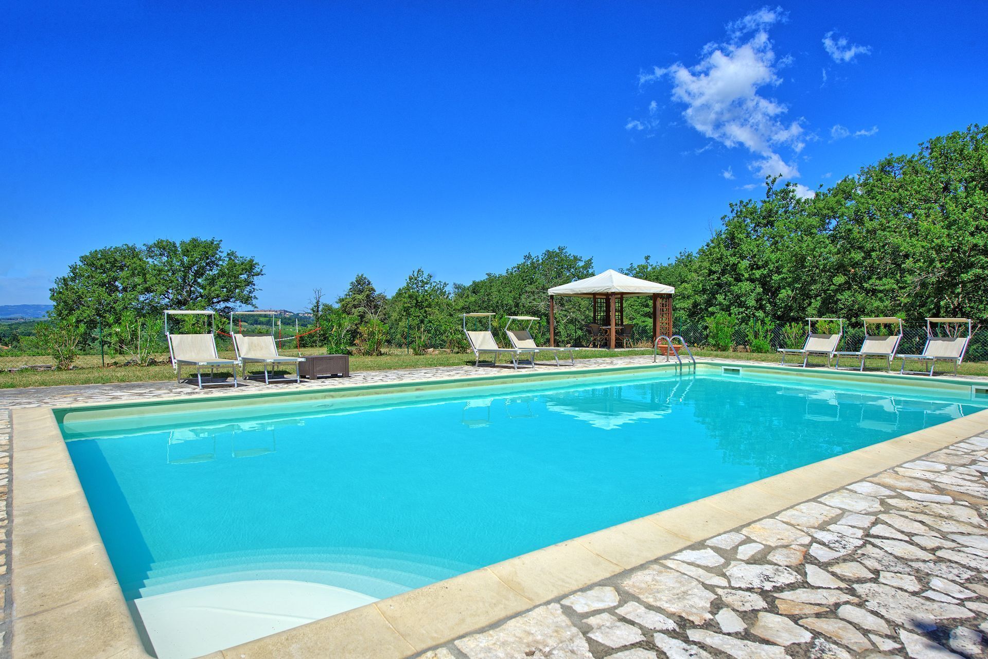 A swimming pool with lounge chairs and a gazebo, surrounded by greenery and a clear sky.