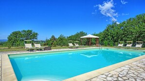 En swimmingpool med solstolar och en pergola, omgiven av grönska och en klar himmel.