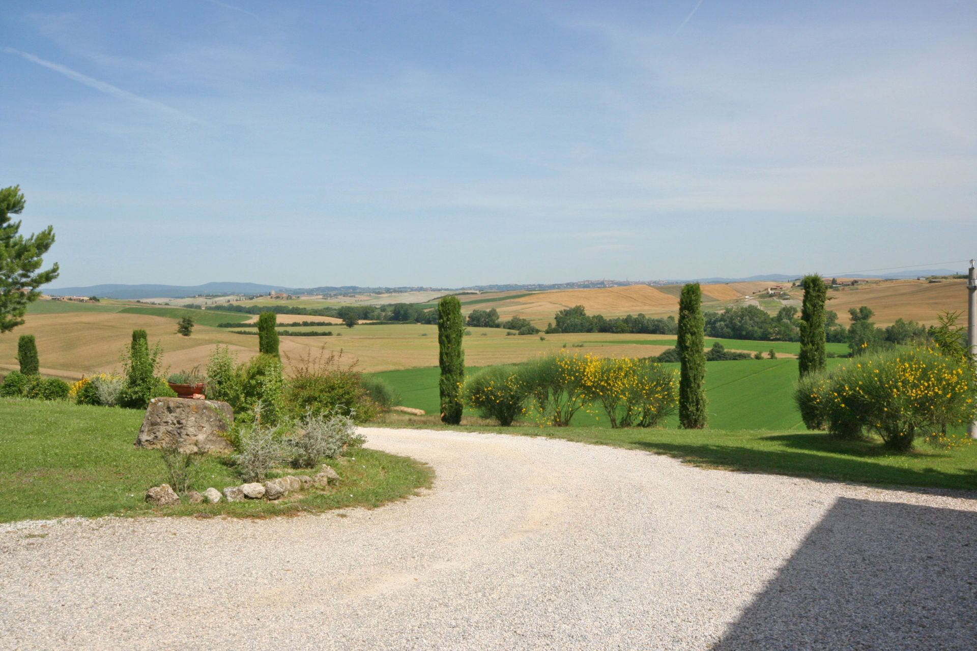 A gravel driveway leading to a rural landscape with rolling hills, green fields, and scattered trees.