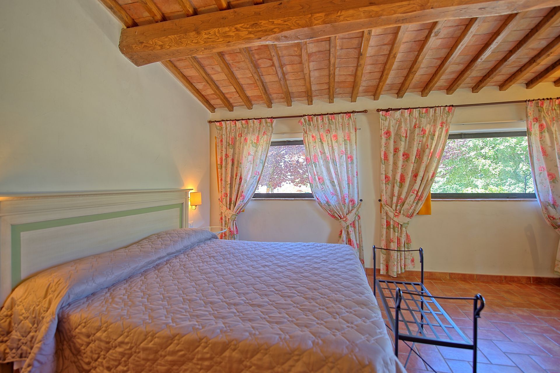 A bedroom with a bed, wooden ceiling beams, floral curtains, and a window with a view of trees.