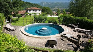 Une piscine ronde entourée de chaises longues et dotée d’une terrasse en pierre, le tout agrémenté de verdure et offrant une vue sur un village et des montagnes en arrière-plan.
