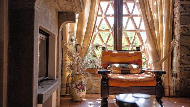 Un intérieur rustique avec une cheminée en pierre, une chaise en bois et une grande fenêtre ornée d’un treillis en bois.