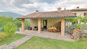 Une terrasse extérieure en pierre, dotée d’un toit en tuiles, entourée de verdure et avec une chaîne de montagnes en toile de fond.