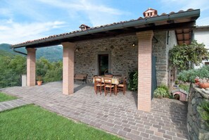 Ein Steinhaus mit überdachter Terrasse, Tisch und Stühlen sowie Blumentöpfen.