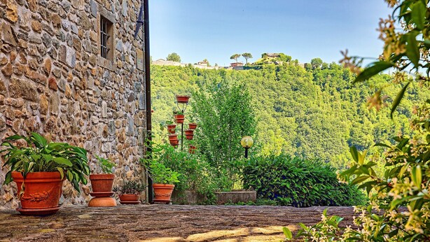 Un bâtiment en pierre, orné de pots en terre cuite, qui offre une vue sur une colline boisée et sur une construction visible au loin.