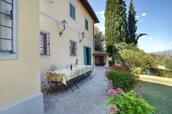 A covered outdoor dining area with a table set for four, surrounded by stone flooring and a garden with various plants and flowers.