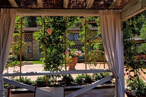 A covered garden area with a pergola, potted plants, and a view of a stone building with a tiled roof.