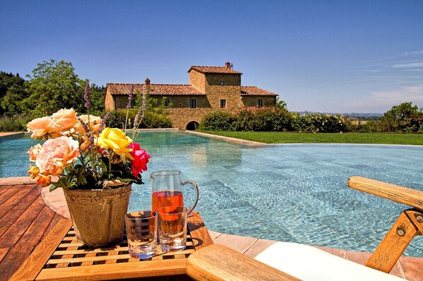 A poolside view with a stone building in the background, a wooden deck, and a table with a vase of flowers and glasses.