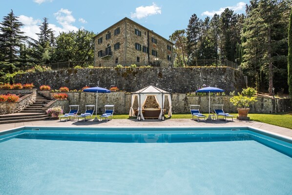 Une piscine avec des chaises longues et des parasols, un bâtiment en pierre doté d’un balcon, et un mur de pierre avec des marches menant à celui-ci.