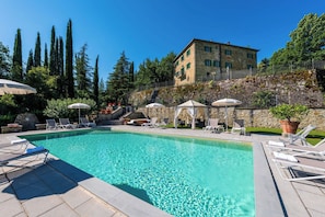 A clear swimming pool with lounge chairs and umbrellas, surrounded by stone walls and greenery.