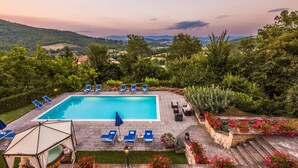 En swimmingpool med liggestole og et pergola-område, omgivet af frodigt grønt og udsigt til fjerne bakker.