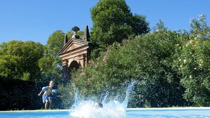 A swimming pool with a person splashing water, surrounded by lush greenery and a historic building in the background.