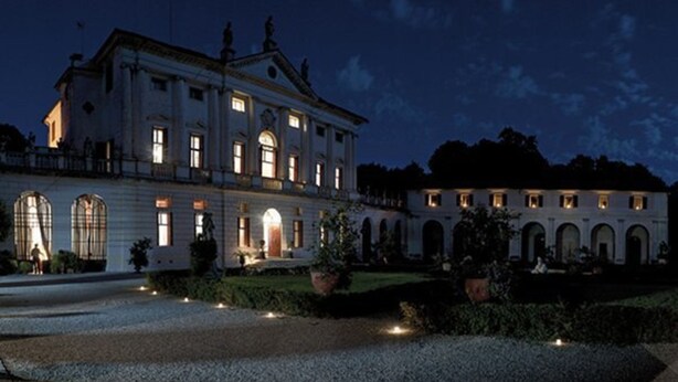 A large, historic building with multiple arched windows and a central entrance, surrounded by a well-maintained garden.