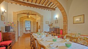 Uma sala de jantar com uma mesa comprida posta para refeição, cadeiras de madeira e um arco de tijolos que leva a outro cômodo.