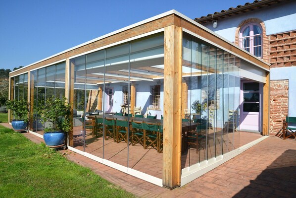 Une terrasse fermée par des baies vitrées, avec une table et des chaises, entourée de plantes en pot et bordée d’un chemin en briques.