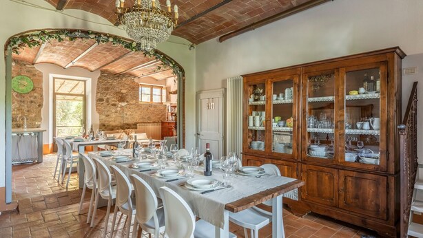 Uma sala de jantar com uma mesa comprida posta para uma refeição, um armário de madeira com portas de vidro e uma parede de pedra ao fundo.