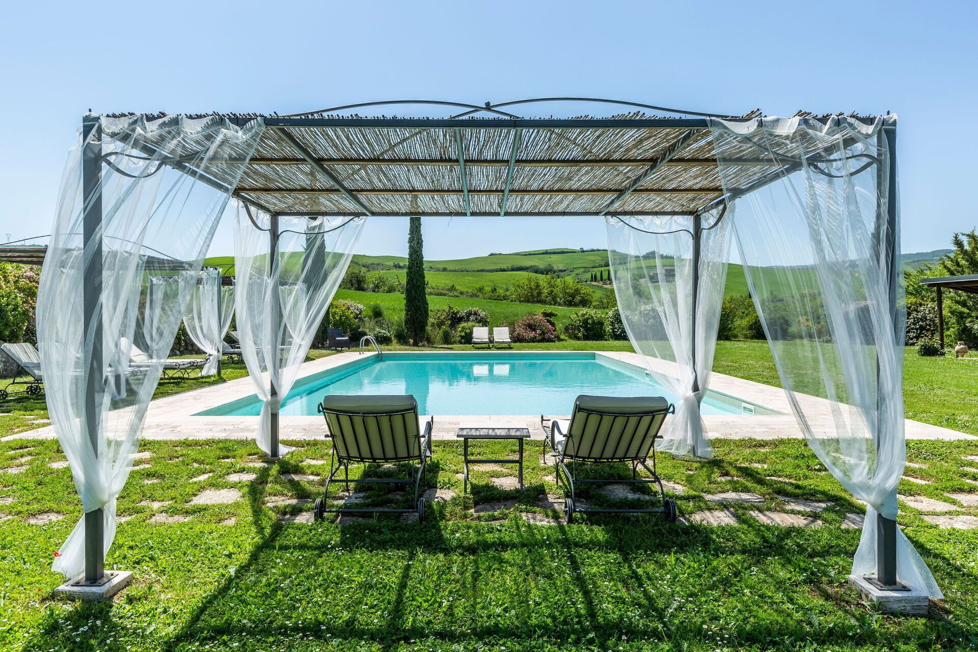 Ein überdachter Poolbereich im Freien mit zwei Liegestühlen und einem Tisch, umgeben von einer Pergola mit luftigen Vorhängen.
