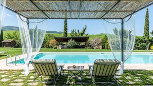 Área de piscina descoberta coberta, com duas cadeiras de descanso, uma mesa e um pergolado com vista para um jardim e colinas.