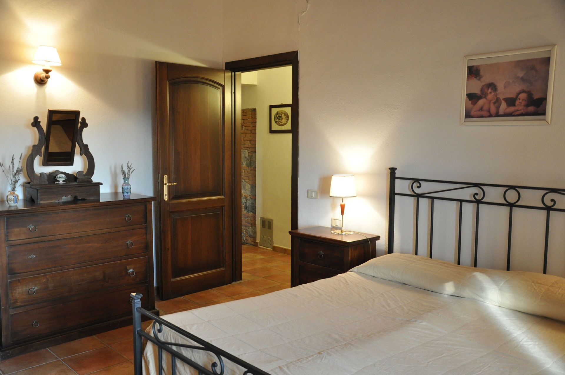 A bedroom with a wooden dresser, a bed with a metal headboard, a nightstand with a lamp, and a door leading to another room.