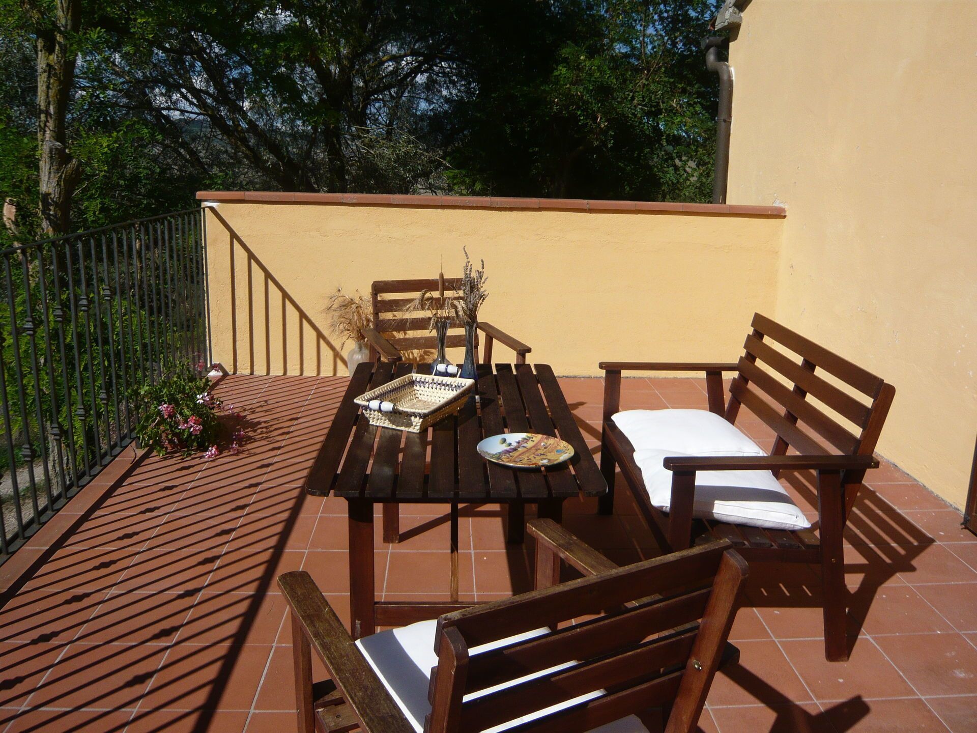 Eine Terrasse mit einem Holztisch und zwei Stühlen, einer Vase mit Trockenpflanzen und einem Metallgeländer.