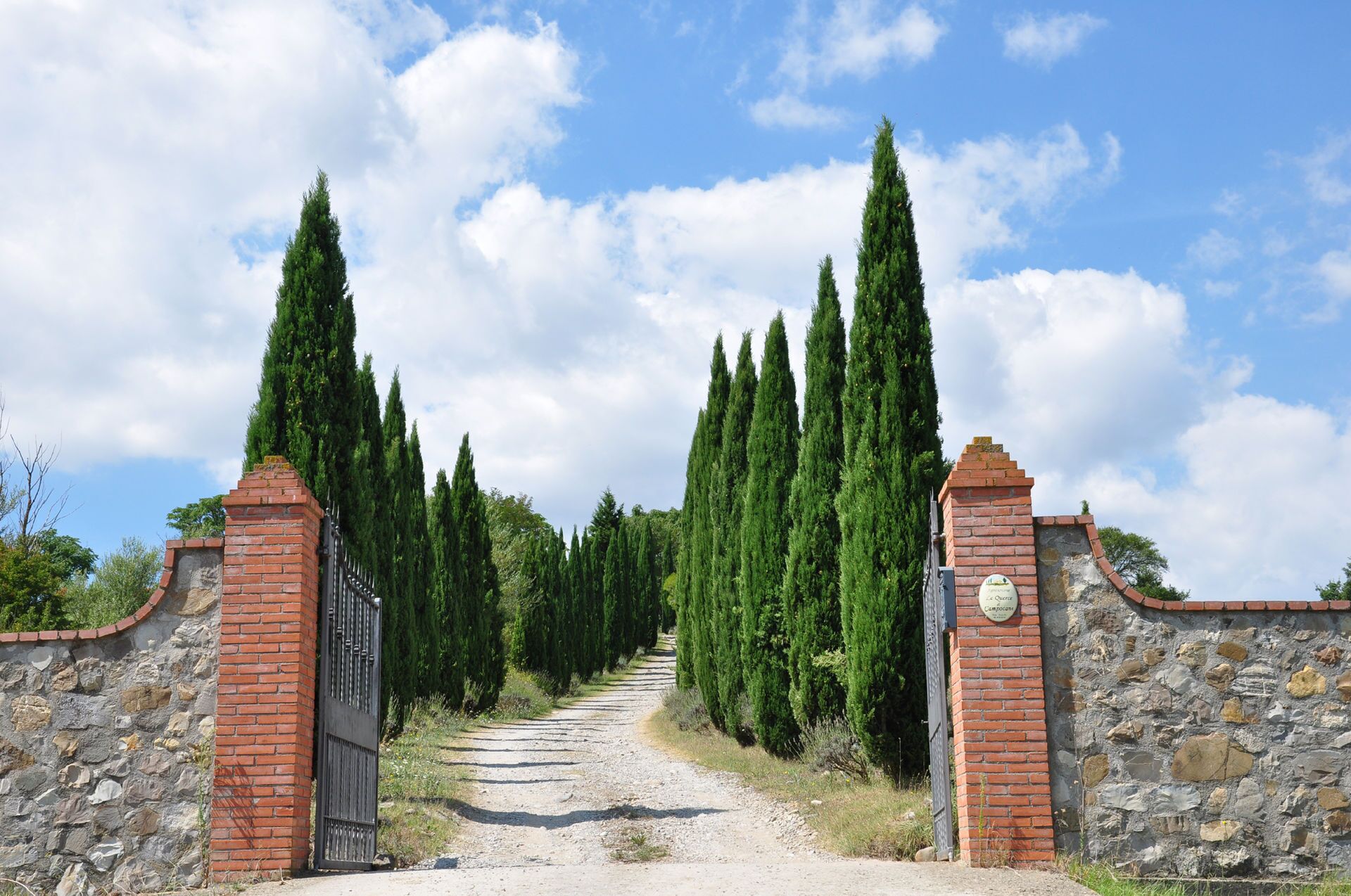 Un sentier qui traverse un portail, de chaque côté duquel s’élèvent de grands cyprès, et qui est bordé de murs en pierre.