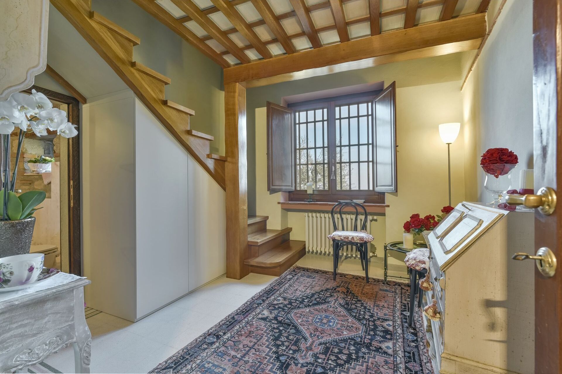 Un couloir avec un escalier en bois, un tapis à motifs, une chaise, une table ornée d’un vase de fleurs et une fenêtre donnant sur un paysage.