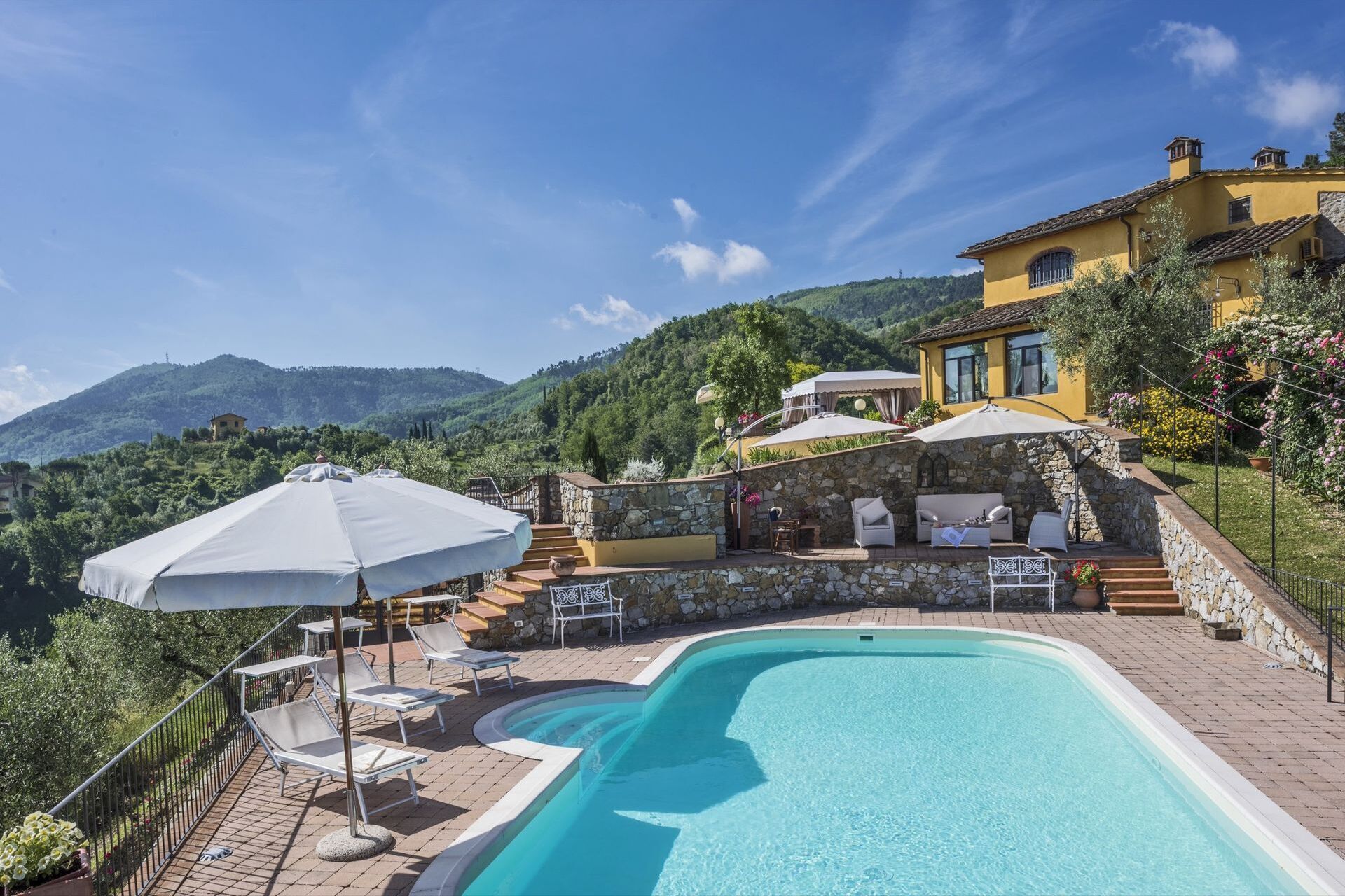 A pool area with lounge chairs and umbrellas, surrounded by a stone wall and a yellow building with a view of the mountains.