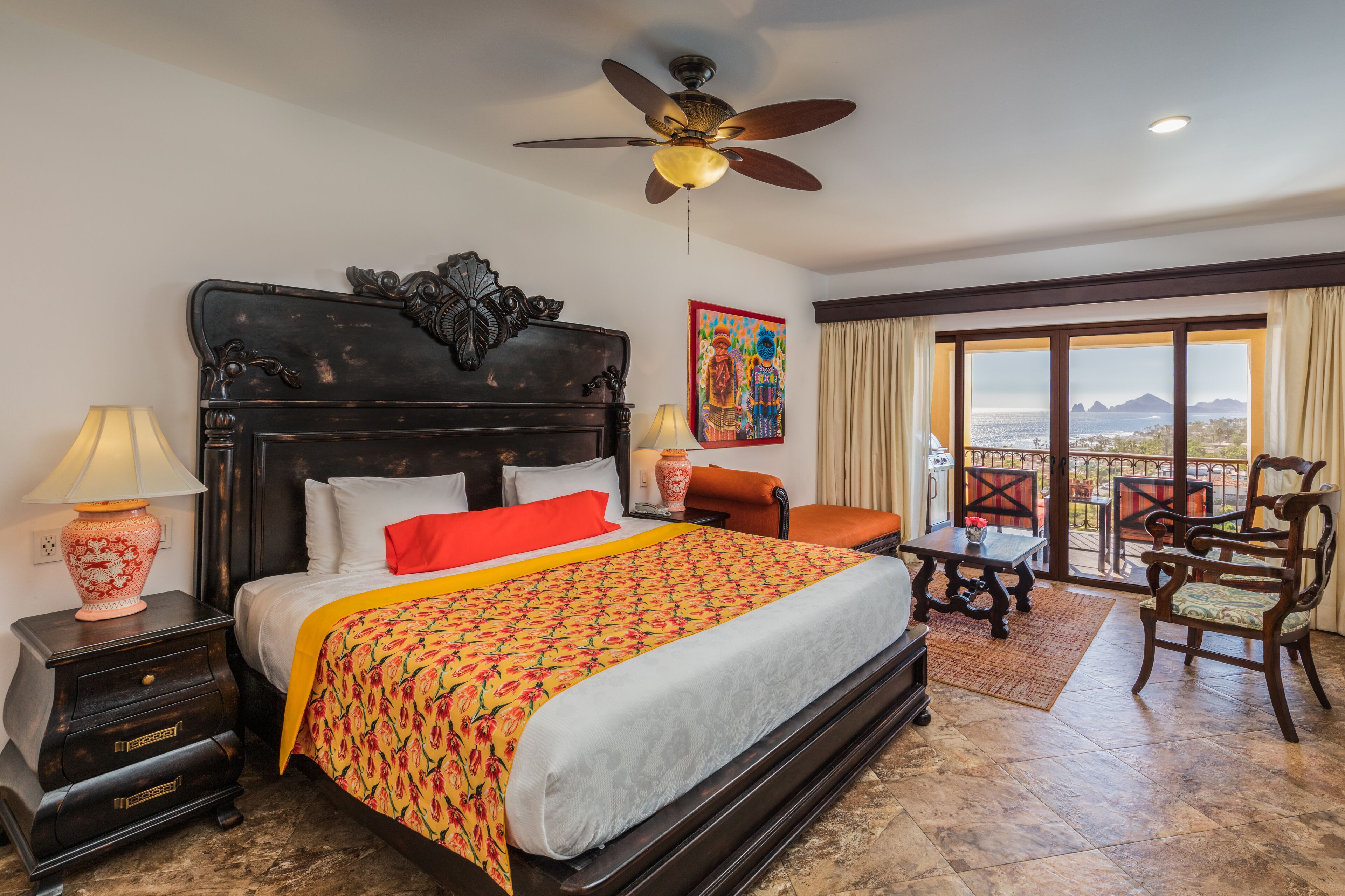 A hotel room with a large bed, a ceiling fan, a painting, a view of the outdoors, and a balcony with chairs.