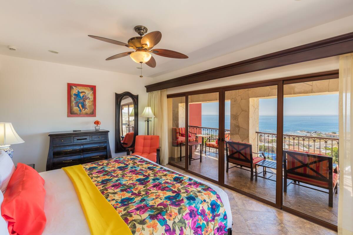 A hotel room with a bed, a ceiling fan, a balcony with chairs, and a view of the ocean.