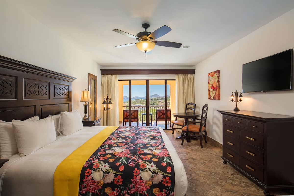 A hotel room with a bed, a ceiling fan, a TV, a balcony with a view, and a dining area.