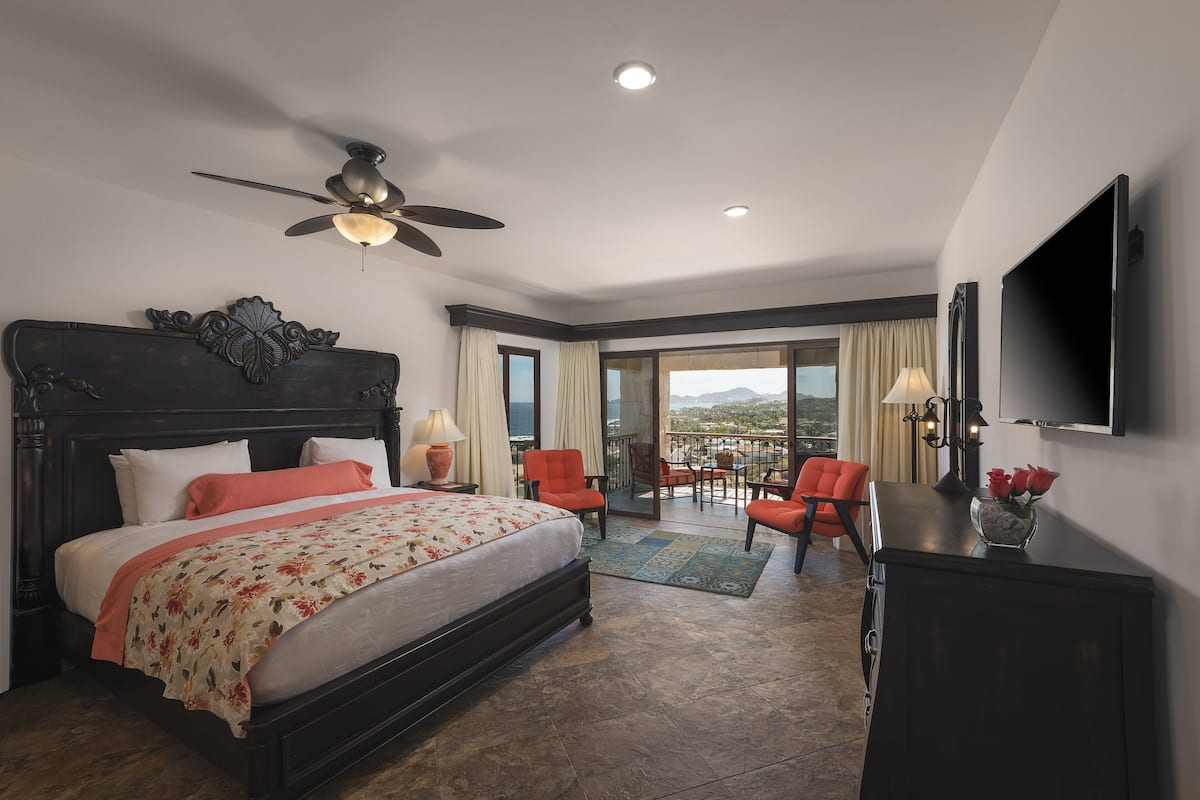 A spacious bedroom with a large bed, a ceiling fan, a TV, and a balcony with chairs and a table.