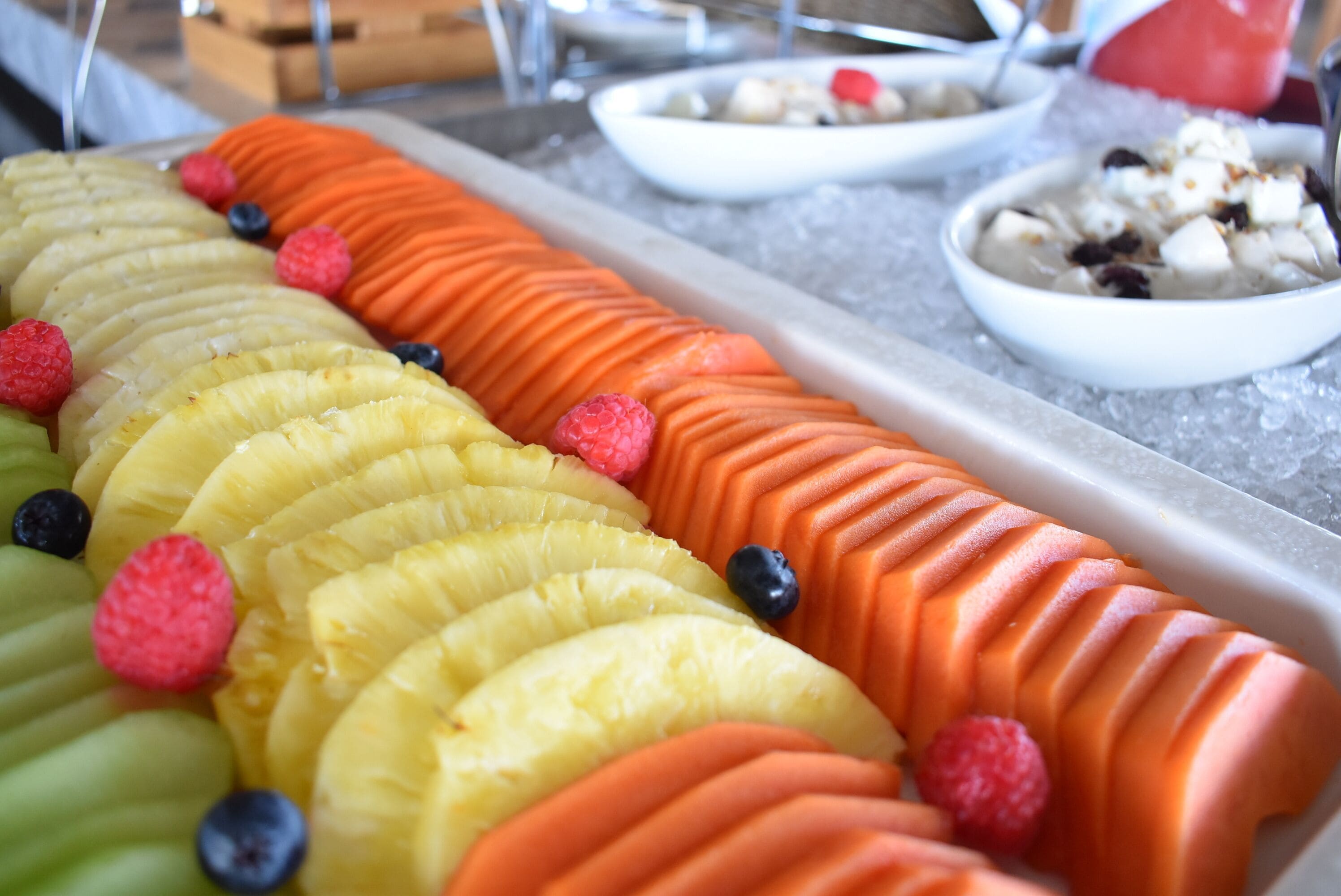 Una bandeja de frutas con piña, plátano y naranja en rodajas, adornada con frambuesas y arándanos.