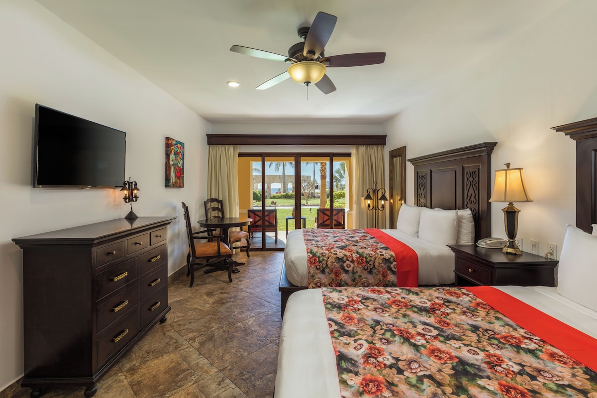 A hotel room with a bed, a desk, a chair, a dresser, a TV, a ceiling fan, and a window with a view of palm trees.