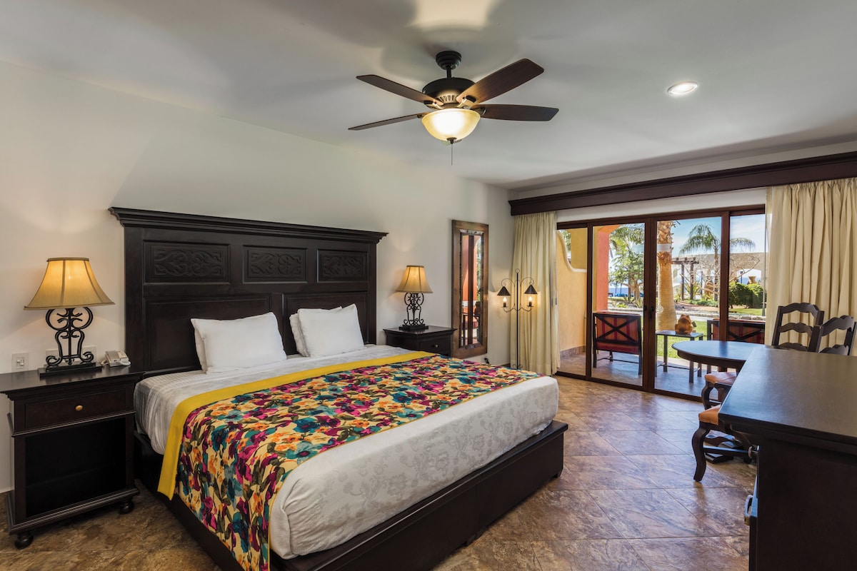 A hotel room with a large bed, a ceiling fan, a desk, and a view of the outdoor patio.