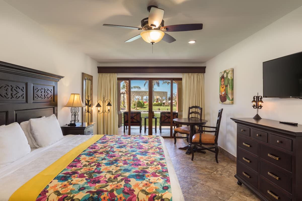 A hotel room with a bed, a ceiling fan, a TV, a desk with a chair, and a view through the window.