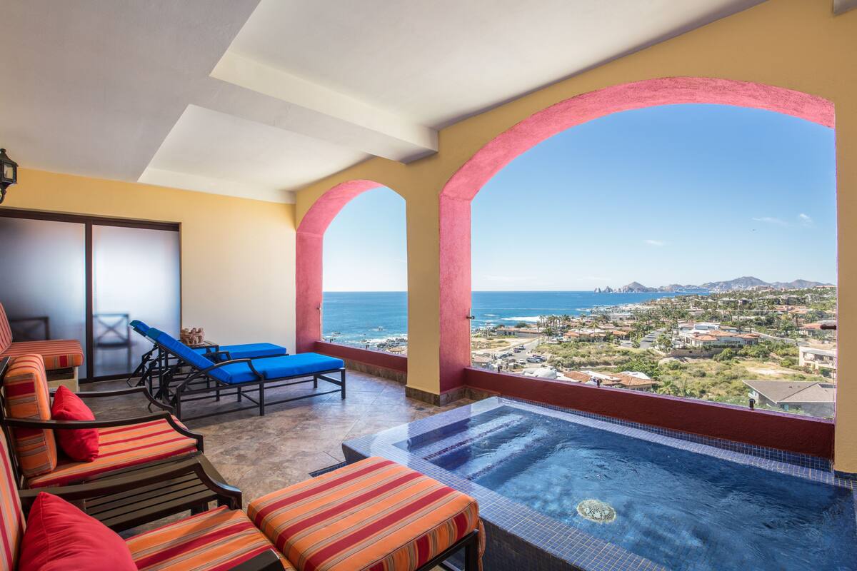 A balcony with a pool, lounge chairs, and a view of the ocean and town.